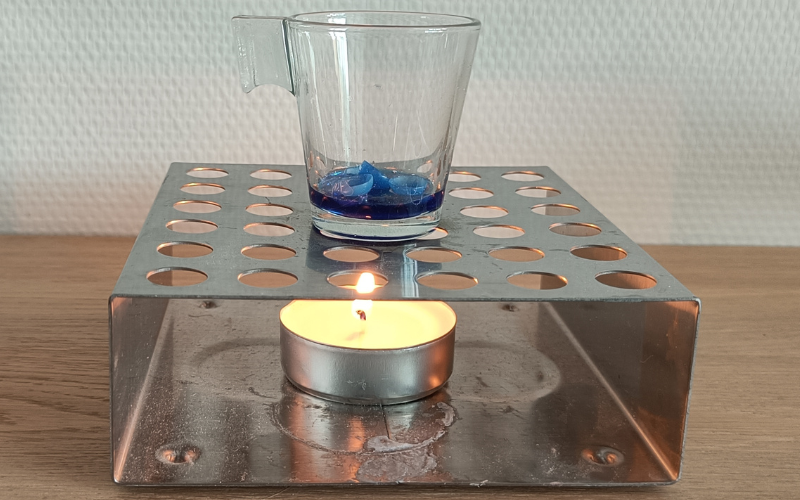 Burning tealight in a large metal holder. On top of tea light holder is a small glass with liquid blue wax and solid blue wax scraps inside