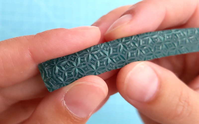 CLose up of hands holding a strip of textured green sheet wax. The surface looks blistered because the top layer is separating