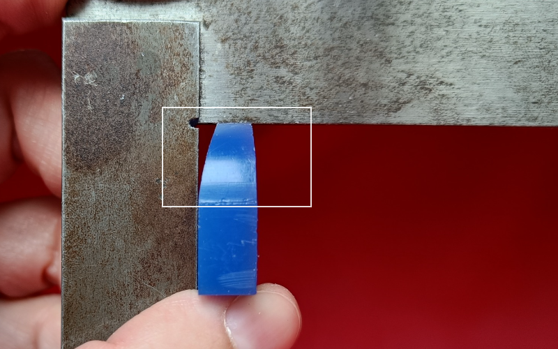Close up of hands holding a piece of blue ring wax against a set square. A white rectangle is drawn around the place you should look at 