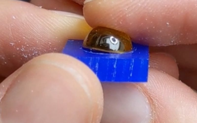 Close up of hands holding a piece of blue wax with a yellow round cabochon on top. The cabochon is partially inside the openiong that's been carved and partially sticking up