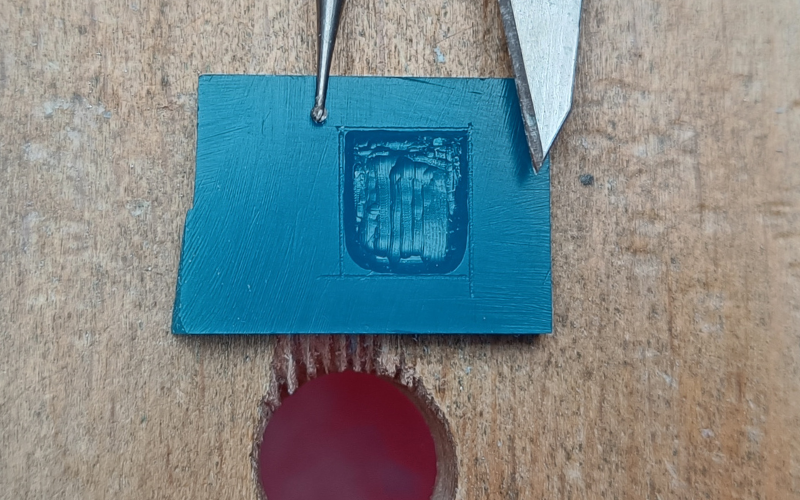 Piece of turquoise wax with a square marked on it. The square is partially burred, the top left corner is burred with a small burr that is lying on the wax. The corner is slightly rounded. The top right corner is done with a stanley knife that is lying on top of the wax. The corner is straight and square