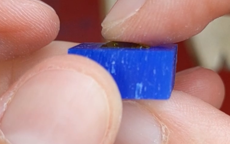 Close up of hands holding a piece of blue wax with a square green stone inside. Girdle of the stone is no longer visible
