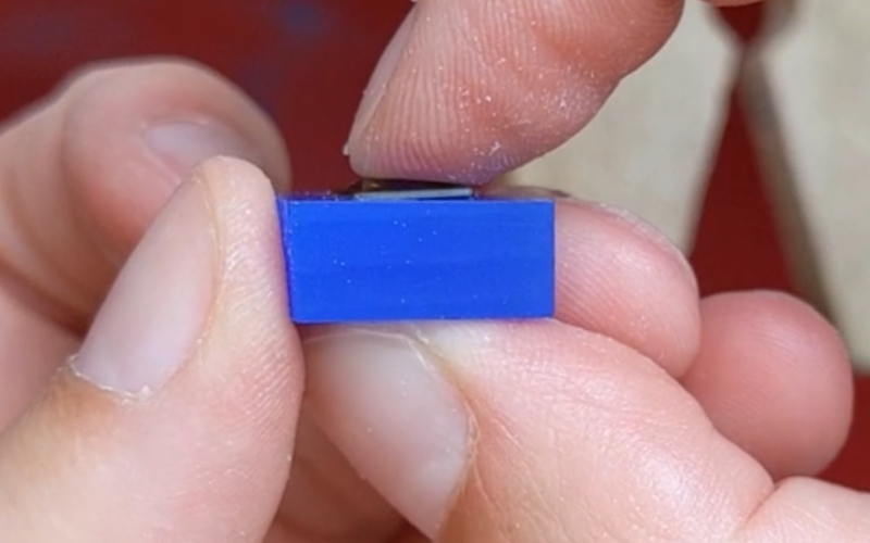 Close up of hands holding a piece of blue wax with a square green stone inside. Girdle of the stone is visible