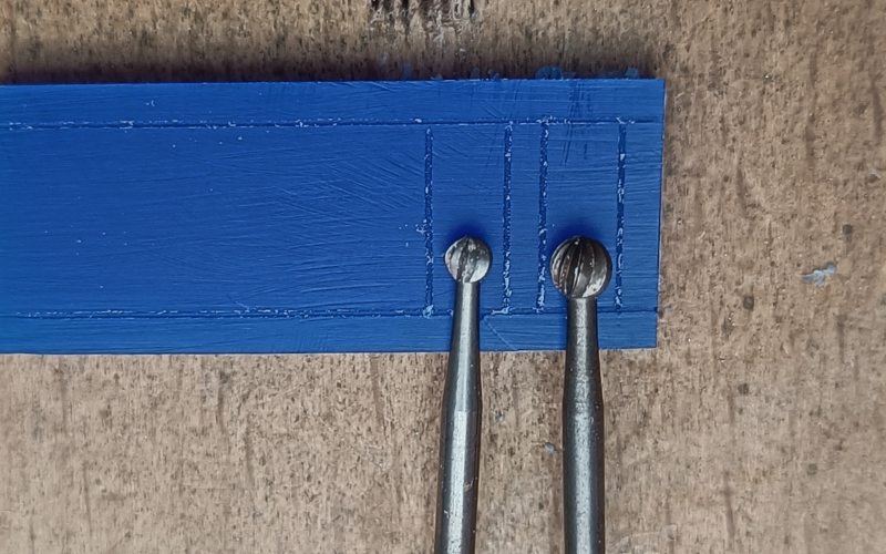 Close up of a piece of blue wax on a bench peg. 2 rectangles are marked on the wax. In one rectangle lies a ball burr that fits nearly perfect between the lines, in the other lies a ball burr that's a lot smaller than the lines. 