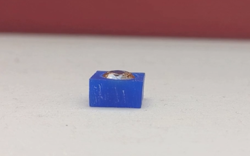Close up of piece of blue wax with a yellow faceted stone inside. The girdle of the stone is inside the wax