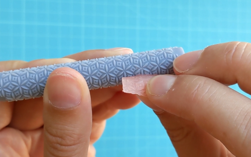 Close up of hands holding a polymer clay texture roller with a piece of pink sheet wax stuck to it, Right hand is carefully trying to peel the sheet wax away from the roller