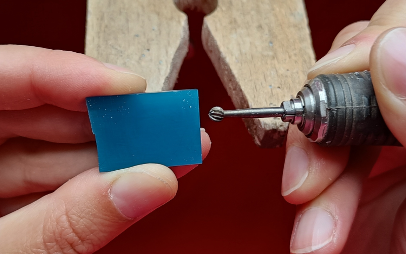 Close up of a hands at the bench. Left hand is resting against the peg holding a piece of turquoise wax, right hand is resting against peg and holding a burr