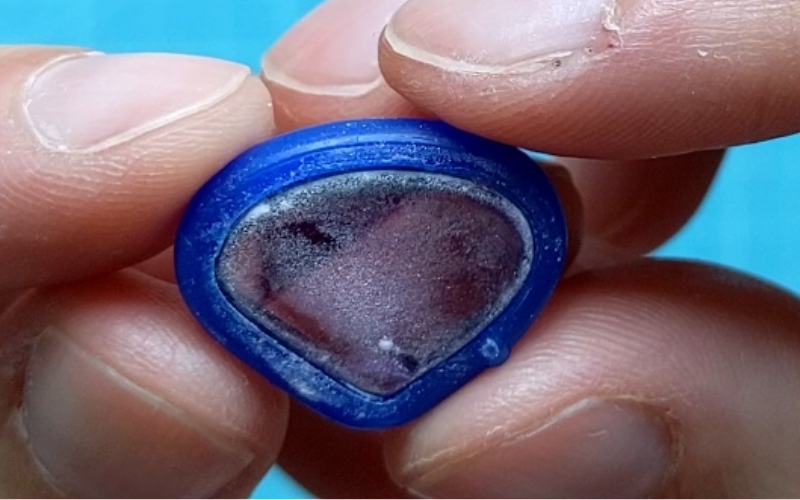 Close up of hands holding a cabochon purple stone with a layer odf talcum powder on the bottom visible. A bezel from blue wax wire fits around the stone
