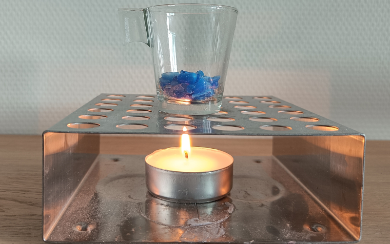 Burning tealight in a large metal holder. On top of tea light holder is a small glass with blue wax pieces inside