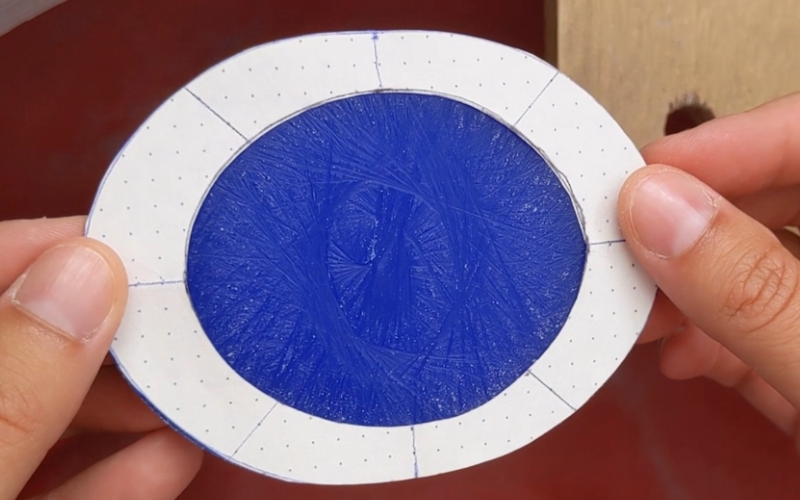 Close up of hands holding an oval piece of blue wax with a paper tamplate on top that has the correct size cut out. 