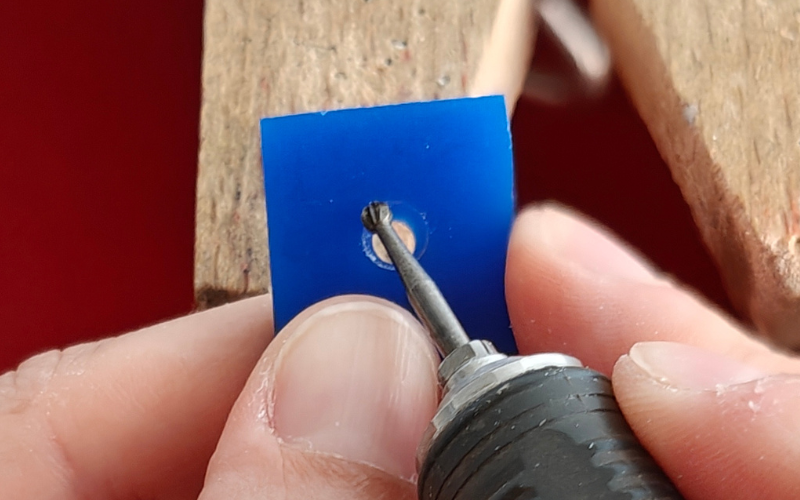 Close up of hands holding a piece of blue square wax with a hole in the center. The piece is angled away from the camera and the other hand is burring the top quarter of the hole. You can't see the bottom quarter and the sides are partially obscured as well 