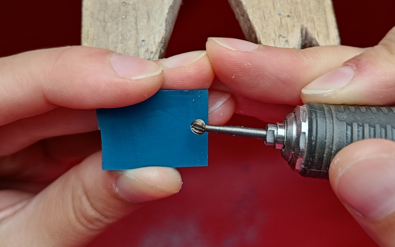 Close up of a hands at the bench. Left hand is resting against the peg holding a piece of turquoise wax, right hand is resting against peg and holding a burr. Fingers of both hands are connected and adding extra support