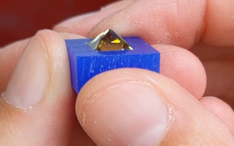 Close up of hands holding a piece of blue wax with a square green stone upside down inside. The pavilion of the stone is in the air, the girdle is inside the wax